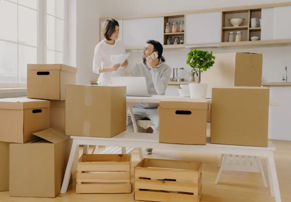 Couple surrounded by cardboard moving boxes in kitchen, man on phone with laptop open